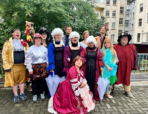 Die Theatergruppe Puzzle posiert mit Ihren Kostümen für das Stück Zauberflöte in einem Innenhof. Einige Personen tragen weiße Mozart-Perücken oder schicke Ballkleider