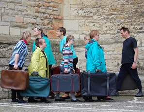 7 Menschen mit Koffern in den Händen laufen im Kreis. Alle schauen geradeaus.